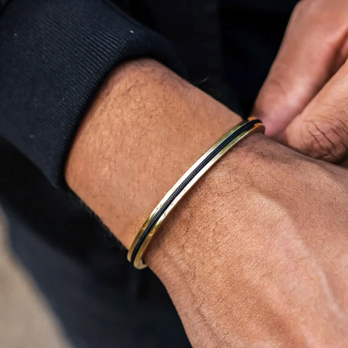 Gold / Black || Man’s wrist featuring a slim gold and black minimalist bracelet with textured center detail against dark sleeve