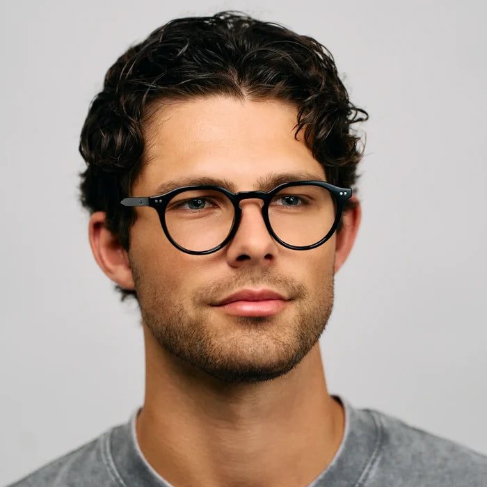 Black || Round prescription glasses with black acetate frame and keyhole bridge worn by man against plain background in natural light